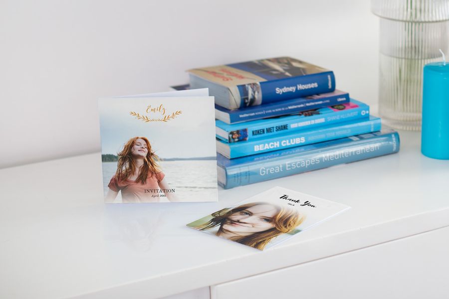 Deux faire-parts de communion personnalisés sur une table blanche avec des livres de voyage bleus en arrière-plan.